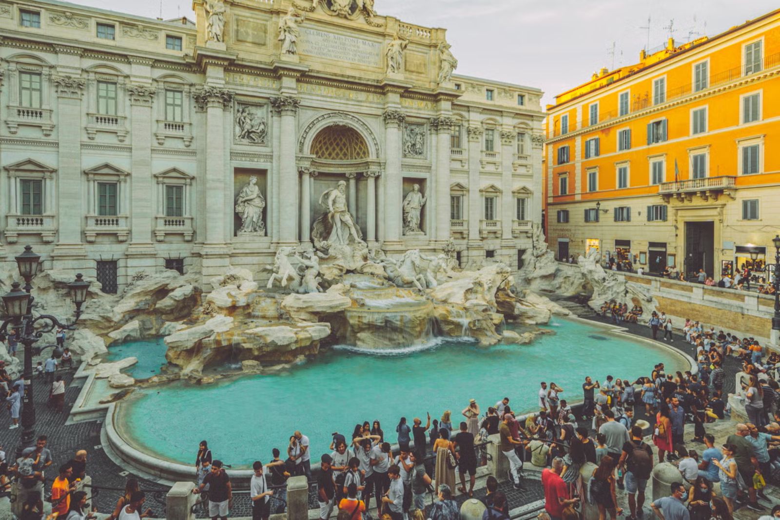The Trevi Fountain in Rome seen from above, its baroque facade and turquoise basin ringed by a dense crowd of tourists