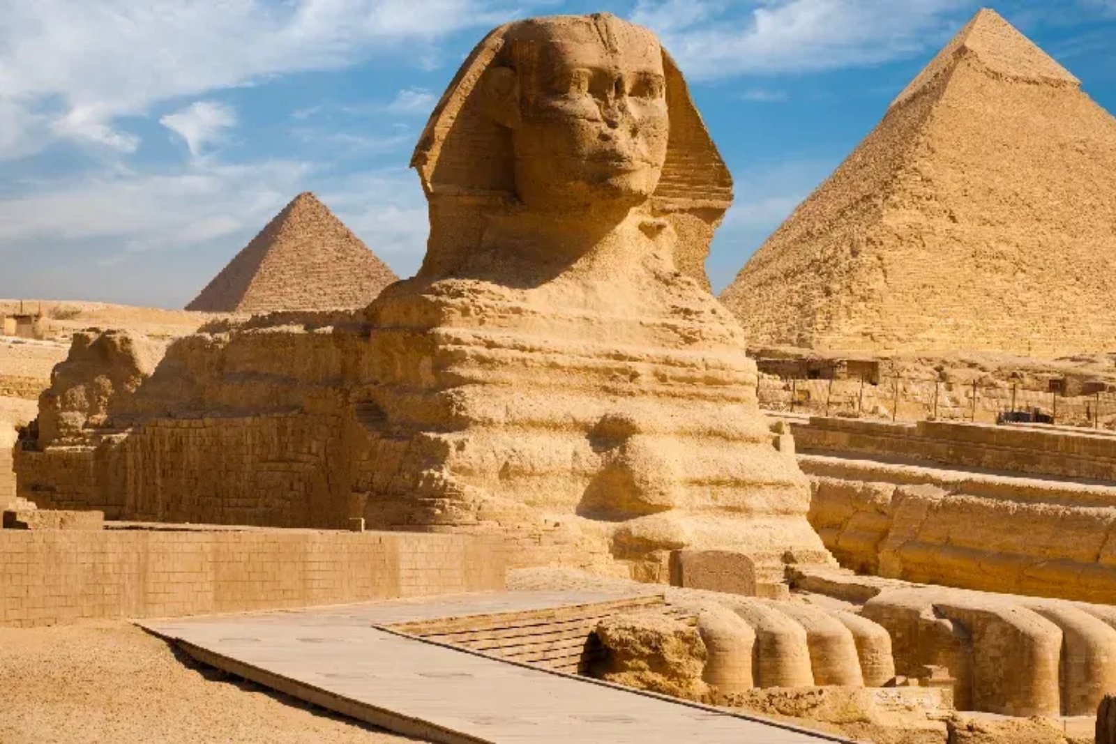 The Great Sphinx of Giza up close, its forepaws in front and two pyramids behind it under a blue sky