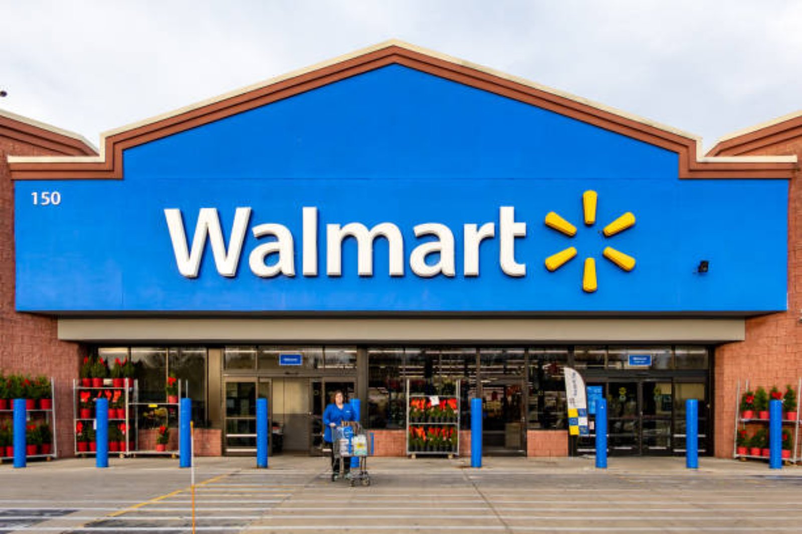 The front of a Walmart store, with its blue sign and yellow spark logo over the entrance and a shopper pushing a cart outside