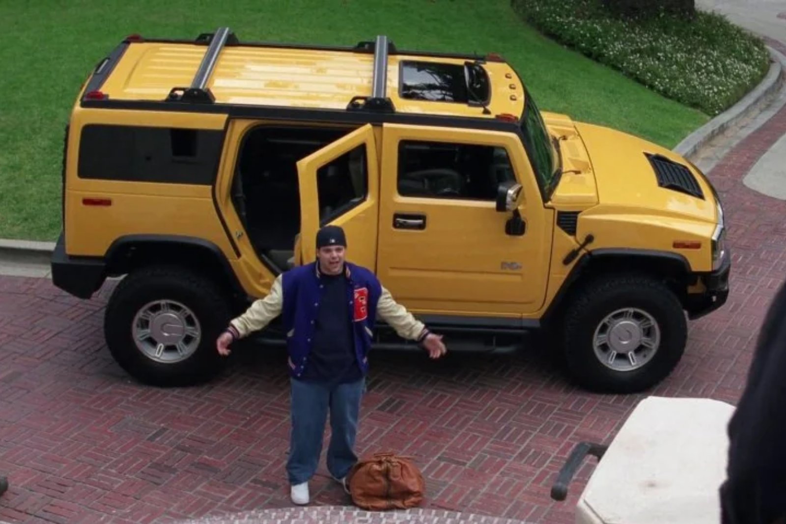 A yellow Hummer H2 on a brick driveway with its back door open, and a man in a varsity jacket standing in front of it with his arms out