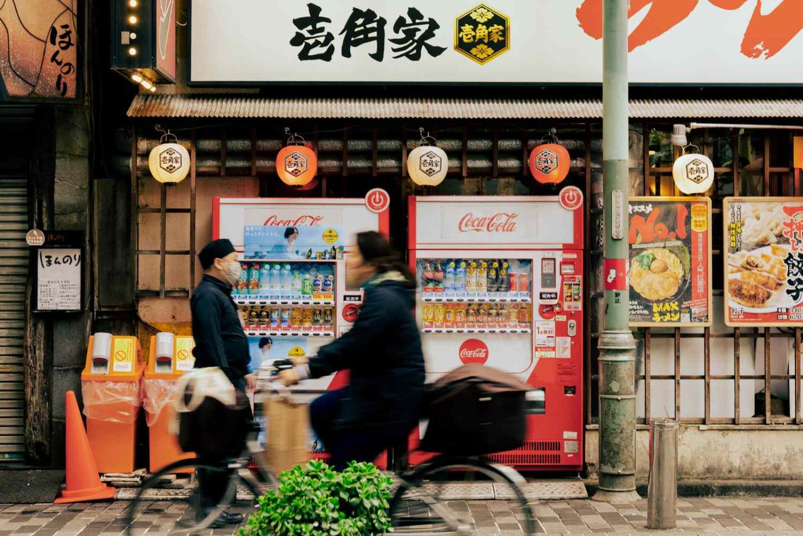 Two red Coca-Cola vending machines outside a ramen shop in Japan, with a cyclist riding past
