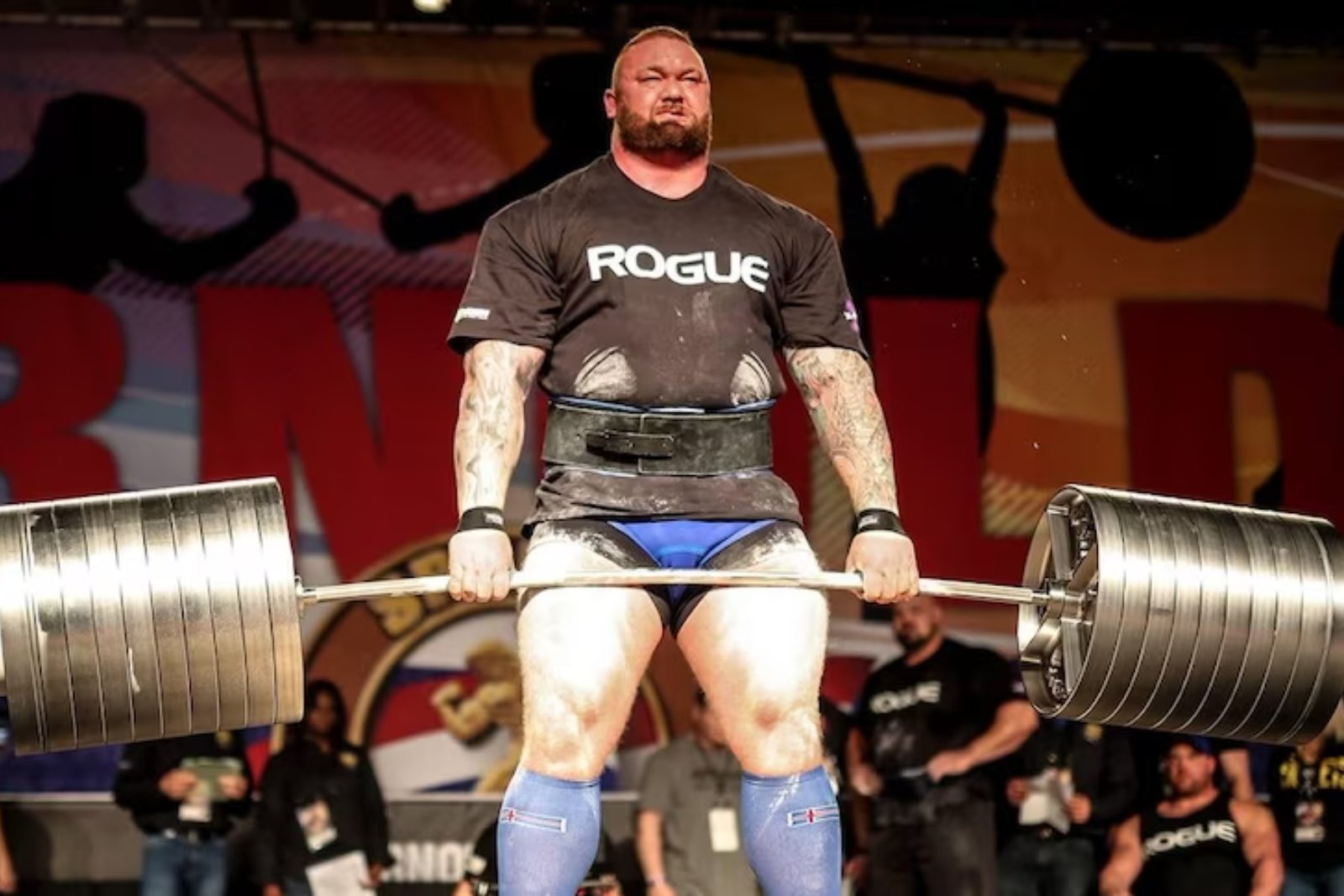 Hafþór Björnsson in a Rogue T-shirt and lifting belt, holding a heavily loaded barbell at the top of a deadlift on a competition stage