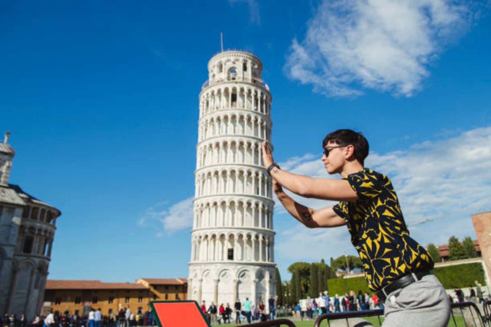 The Leaning Tower of Pisa tilting against a blue sky, its stacked white levels of arches behind a tourist posing as if holding it up