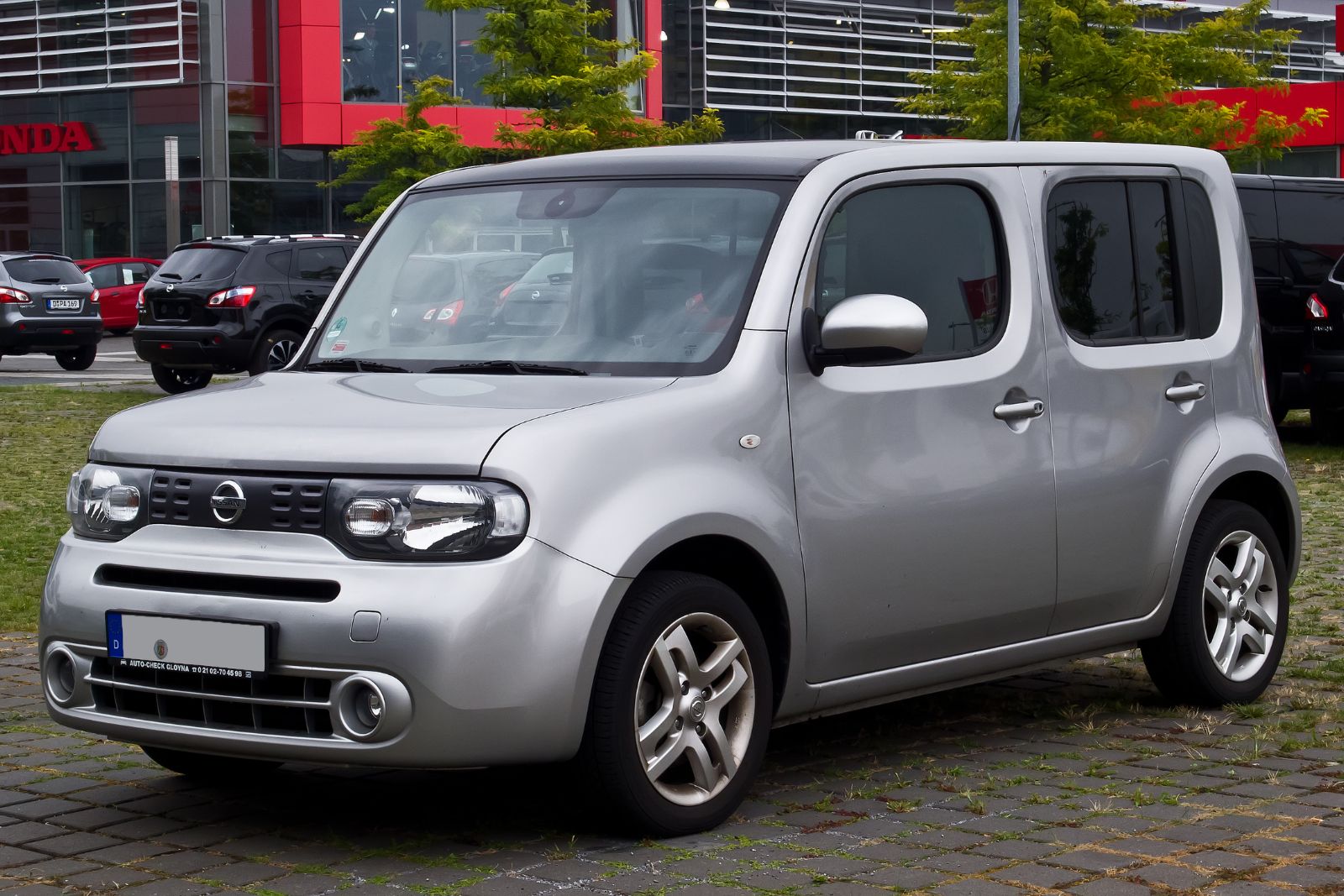 A silver Nissan Cube parked outside a car dealership, seen from the front left, showing its tall boxy body