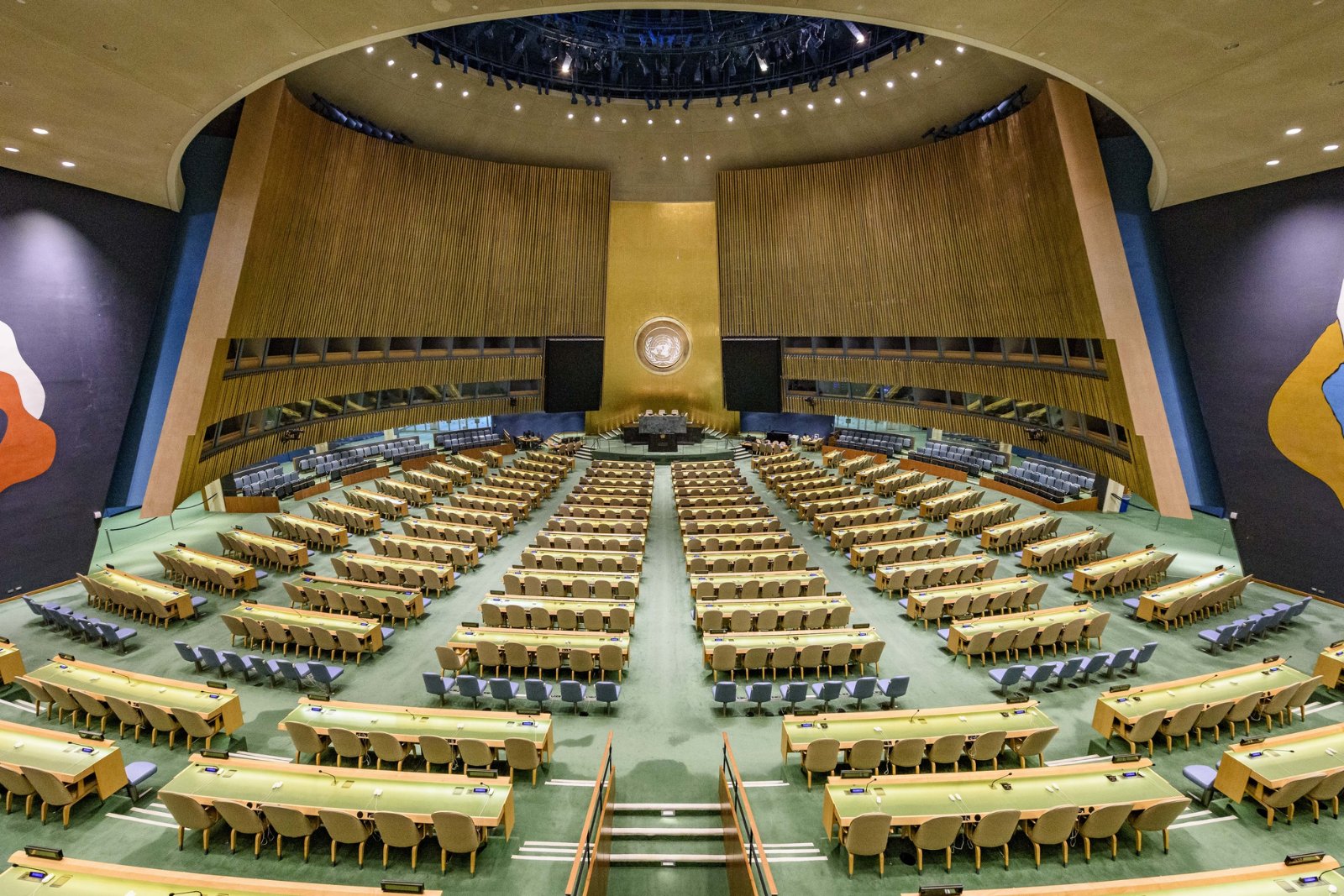 The empty United Nations General Assembly Hall in New York, rows of green desks facing the gold wall behind the podium.