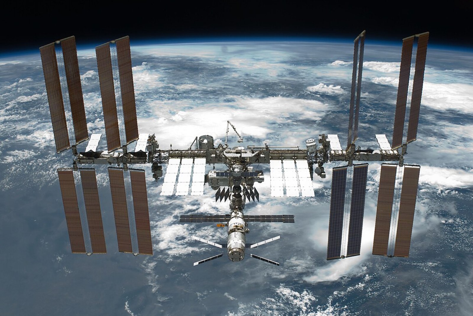 The International Space Station in orbit, its long truss and solar array wings spread wide, with the cloud-covered curve of Earth behind it